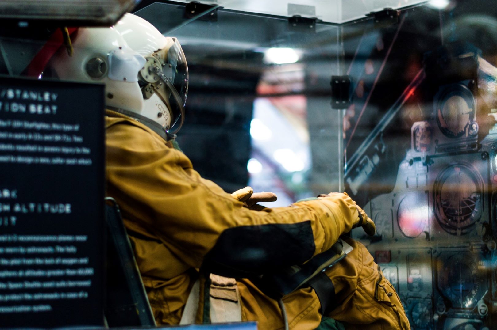 Person in a yellow flight suit and helmet sitting in a cockpit, facing the control panel in an aviation or space setting